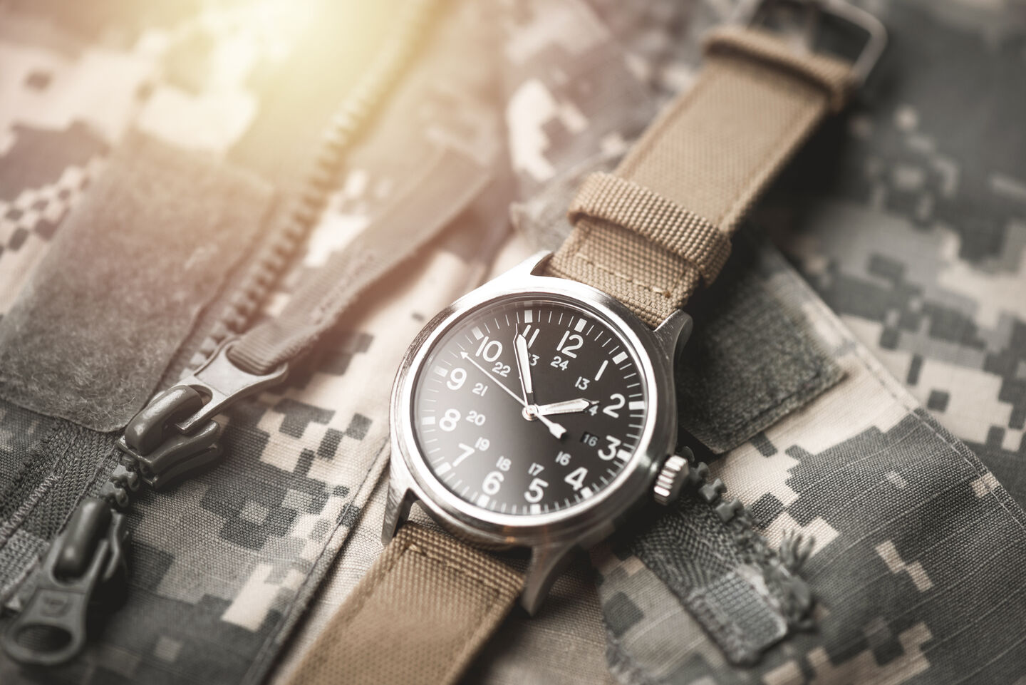 A close-up of a silver, chunky wristwatch on top of a camouflage jacket. A light source glints, reflecting off the metal surface of the watch.